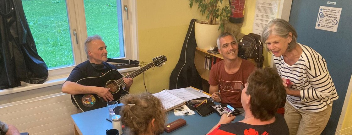 Sechs Personen, eine davon im Rollstuhl, sitzen in einer Wärmestube an einem Tisch beisammen, während ein Mann Gitarre spielt.