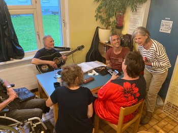 Sechs Personen, eine davon im Rollstuhl, sitzen in einer Wärmestube an einem Tisch beisammen, während ein Mann Gitarre spielt.