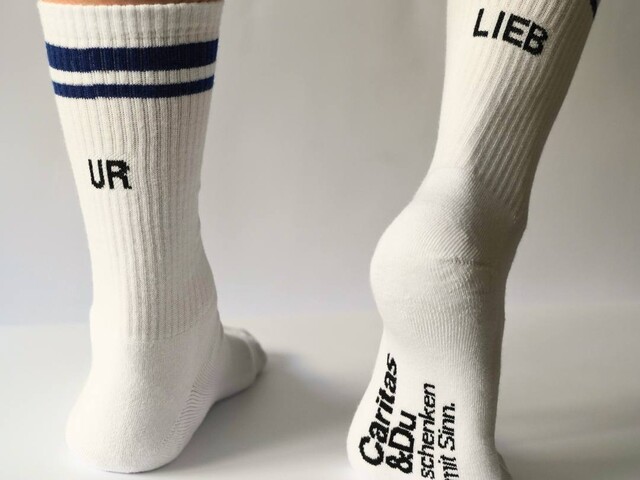Weiße Tennissocken mit der Aufschrift "UR LIEB" und dem offiziellen Caritas Logo.