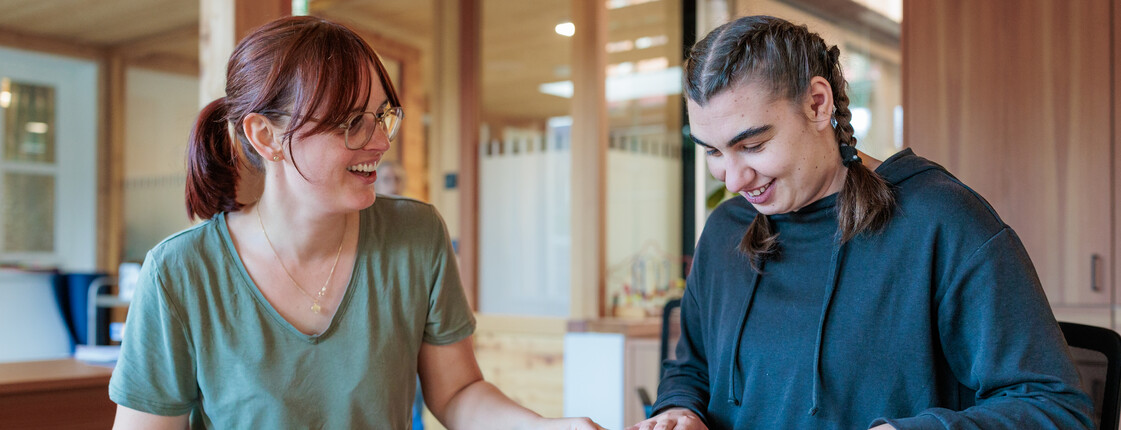 Eine Caritas Mitarbeiterin und eine Frau mit Beeinträchtigung sitzen an einem Tisch in einem Wintergarten und spielen das Gesellschaftsspiel Memory.