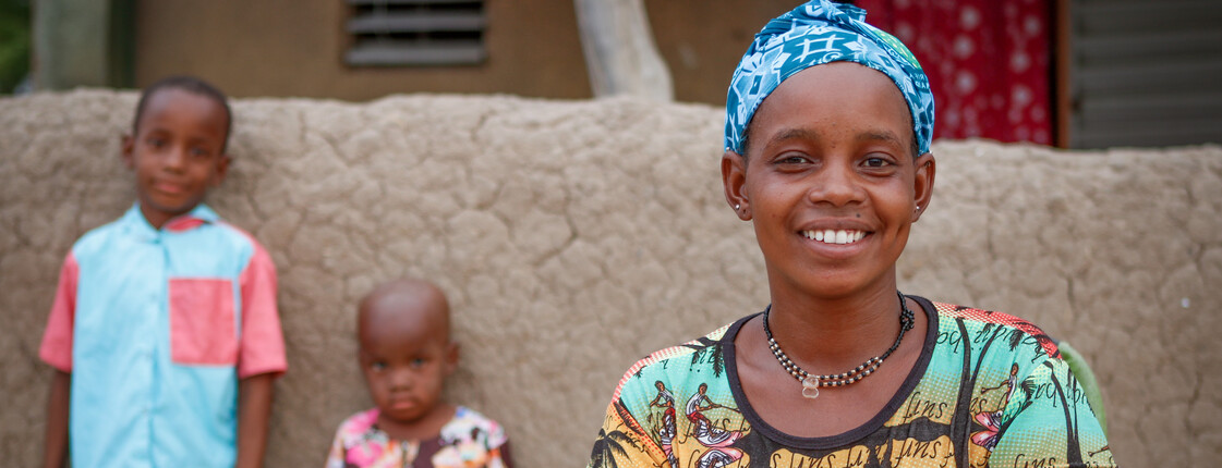 Portrait einer afrikanischen Frau mit bunter Kleidung. Im Hintergrund lehnen zwei Kinder mit dem Rücken an einer Lehmwand.