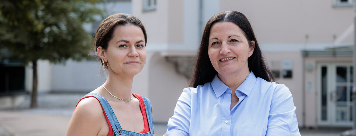 Portrait zwei Frauen in einem Innenhof. Die Frau rechts hat die Arme vor dem Körper verschränkt.