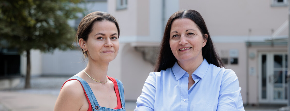 Portrait zwei Frauen in einem Innenhof. Die Frau rechts hat die Arme vor dem Körper verschränkt.