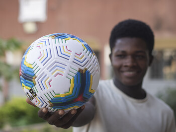 Nahaufnahme eines weißen Fußballs mit bunten Streifen, der von einem lächelnden Jugendlichen vor die Kamera gehalten wird.