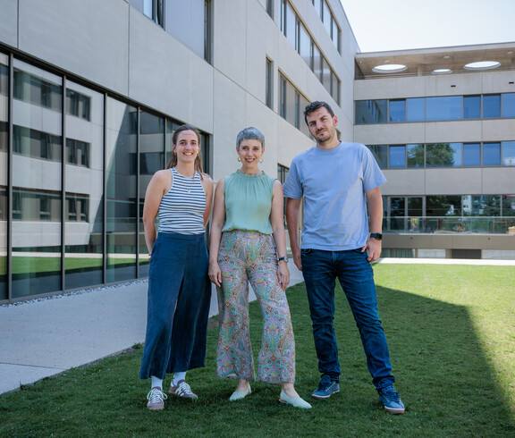 Drei Personen stehen auf einer grünen Wiese im Innenhof eines U-förmigen, modernen Gebäudes.