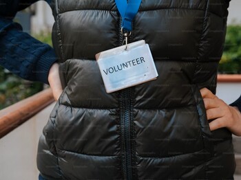Eine Frau hat eine schwarze Steppweste an und trägt ein Schild mit der Aufschrift "Volunteer".