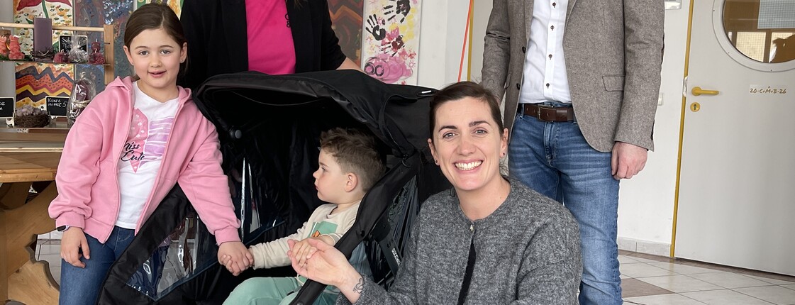 Drei Personen und ein Kind stehen in einem Foyer um einen Kinderwagen, in dem ein Kleinkind sitzt, zu einem Gruppenfoto zusammen.