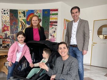 Drei Personen und ein Kind stehen in einem Foyer um einen Kinderwagen, in dem ein Kleinkind sitzt, zu einem Gruppenfoto zusammen.