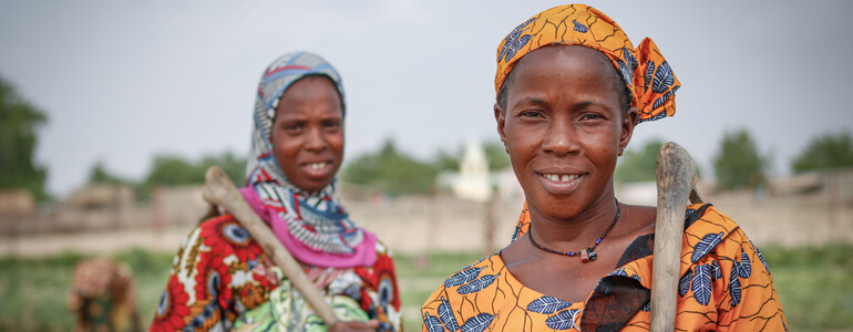 Portrait zweier Frauen in tradioneller afrikanischer Kleidung, die jeweils eine Feldhacke aus Holz geschultert haben.