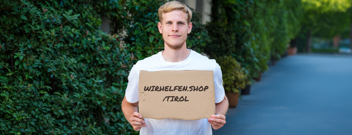 Ein Mann, der ein weißes T-Shirt trägt, hält ein Kartonschild mit der Aufschrift "wirhelfen.shop/tirol" in die Kamera.