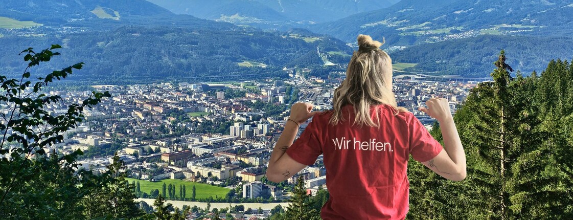 Eine Frau, die ein rotes T-Shirt mit der Aufschrift "Wir helfen" trägt, steht vor einem Innsbrucker Panorama.