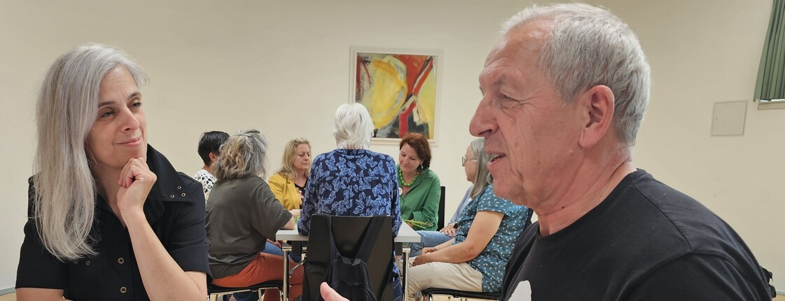 Ein Mann und eine Frau sitzen während eines Workshops an einem Tisch, auf dem ein selbstgemachtes Plakat liegt, und unterhalten sich.