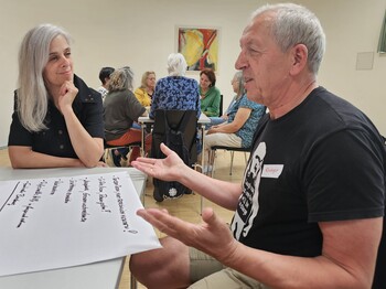 Ein Mann und eine Frau sitzen während eines Workshops an einem Tisch, auf dem ein selbstgemachtes Plakat liegt, und unterhalten sich.