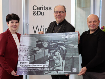 Drei Personen stehen vor einem Caritas Roll-Up und präsentieren ein Plakat mit einer schwaz-weiß Fotografie.