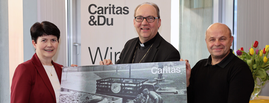 Drei Personen stehen vor einem Caritas Roll-Up und präsentieren ein Plakat mit einer schwaz-weiß Fotografie.