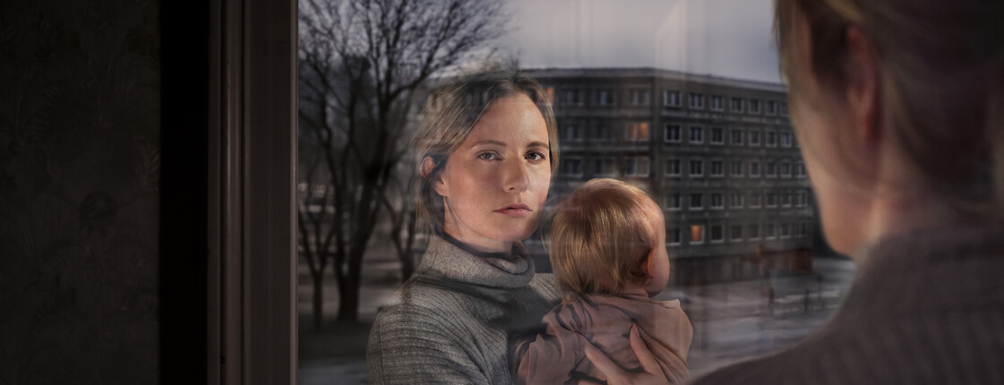 Eine Frau mit Kind am Arm schaut aus dem Fenster. Ihr Gesicht spiegelt sich im Fensterglas wider. Vor dem Fenster ist eine Wohngegend erkennbar.