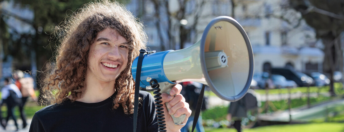 Portrait eines lachenden, jungen Mannes, der bei einer Veranstaltung in ein Megafon spricht.