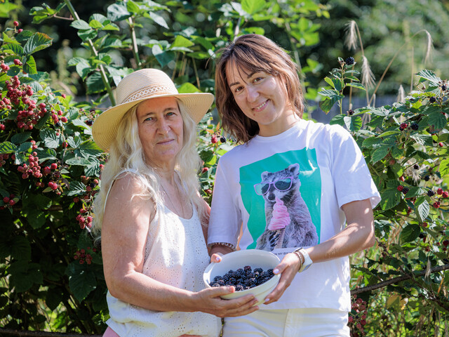 Zwei Frauen mittleren Alters in Sommerbekleidung stehen in einem Garten und halten zusammen eine Schüssel Brombeeren.