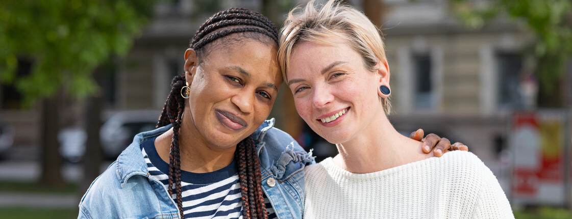 Zwei junge Frauen, die sich im Buddy-Projekt kennengelernt haben, stehen Arm in Arm in einem Park und schauen lachend in die Kamera.