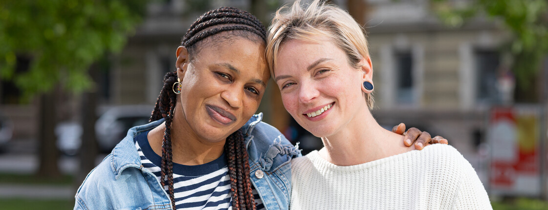 Zwei junge Frauen, die sich im Buddy-Projekt kennengelernt haben, stehen Arm in Arm in einem Park und schauen lachend in die Kamera.