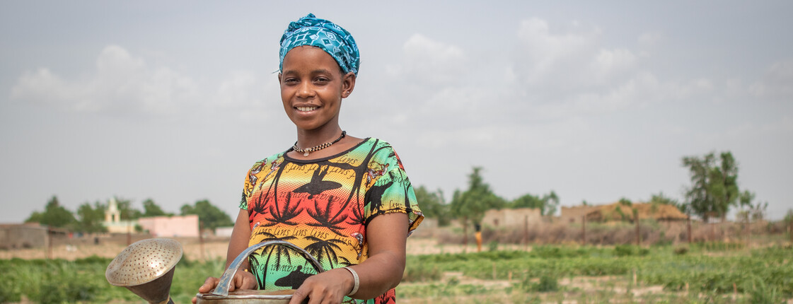 Portrait einer bunt gekleideten Frau in Burkina Faso, die mit einer eisernen Gießkanne an einem Bewässerungsbrunnen auf einem Anbaufeld steht.