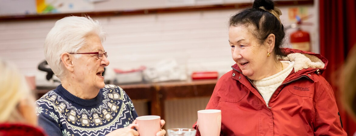 Vier Frauen sitzen an einem gedeckten Küchentisch. Sie trinken Kaffee und unterhalten sich.