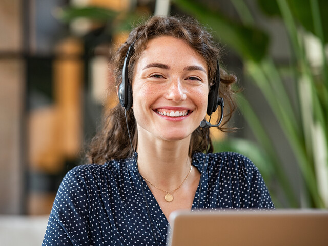 Portrait einer lächelnden jungen Frau, welche Businesskleidung und ein Headset trägt.