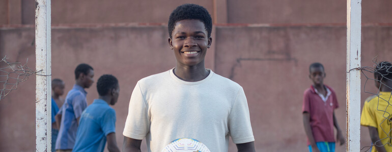 Portrait eines Jugendlichen, der ein weißes T-Shirt trägt und einen Fußball in den Händen hält. Im Hintergrund ist ein Fußballfeld auf dem Kinder Fußball spielen.