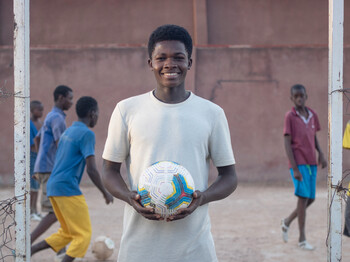 Portrait eines Jugendlichen, der ein weißes T-Shirt trägt und einen Fußball in den Händen hält. Im Hintergrund ist ein Fußballfeld auf dem Kinder Fußball spielen.