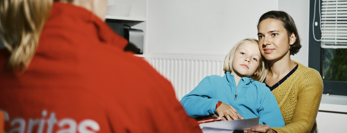Eine Caritas Mitarbeiterin führt an einem Küchentisch ein Beratungsgespräch mit einer Frau, die ein Kind auf dem Schoß hat.