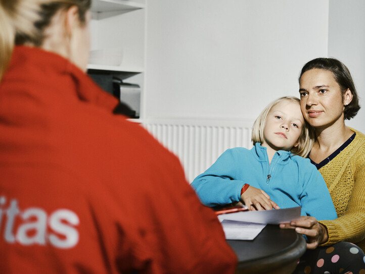 Eine Caritas Mitarbeiterin führt an einem Küchentisch ein Beratungsgespräch mit einer Frau, die ein Kind auf dem Schoß hat.