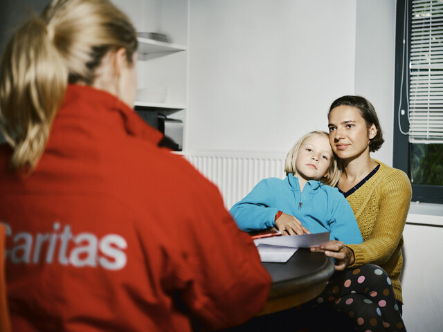 Eine Caritas Mitarbeiterin führt an einem Küchentisch ein Beratungsgespräch mit einer Frau, die ein Kind auf dem Schoß hat.