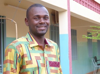 Didier Ouedraogo zu Gast im Welthaus  Portrait eines Mannes aus Burkina Faso, der ein buntes Hemd trägt, und vor einem Hauseingang steht.