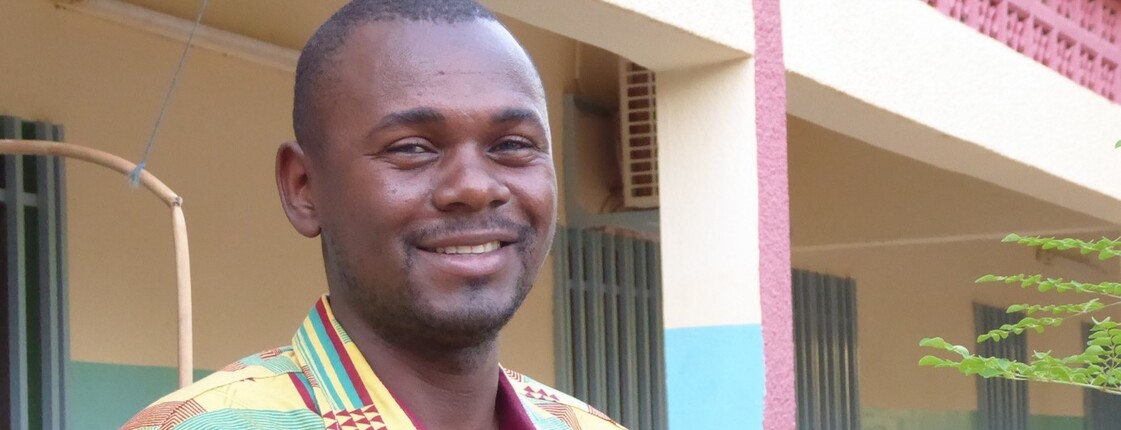 Didier Ouedraogo zu Gast im Welthaus  Portrait eines Mannes aus Burkina Faso, der ein buntes Hemd trägt, und vor einem Hauseingang steht.