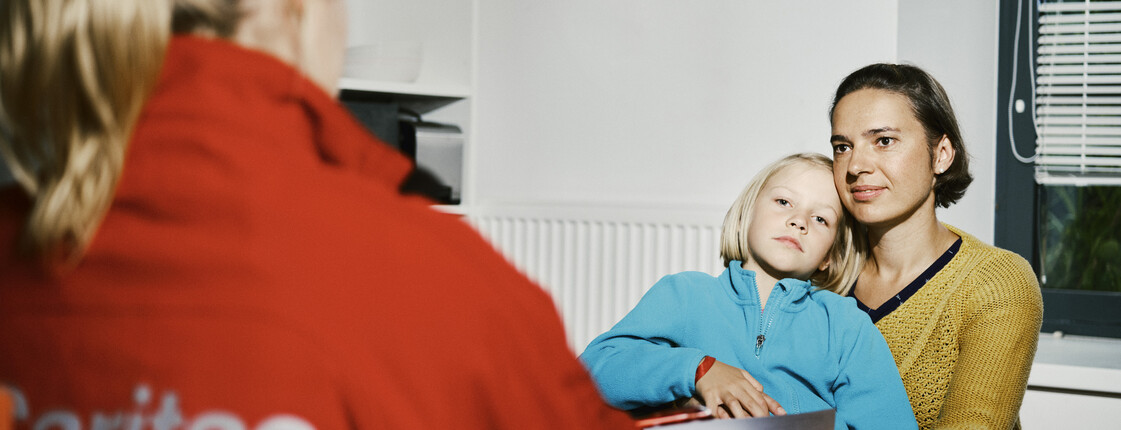 Eine Caritas Mitarbeiterin führt an einem Küchentisch ein Beratungsgespräch mit einer Frau, die ein Kind auf dem Schoß hat.