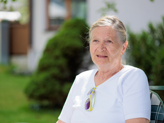 Eine Seniorin mit weißem T-Shirt sitzt im Sommer in einem grünen Garten.