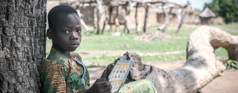 Portrait eines Jungen in traditioneller Kleidung in Burkina Faso, der mit einem Schulheft in der Hand im Schatten unter einem Baum sitzt..