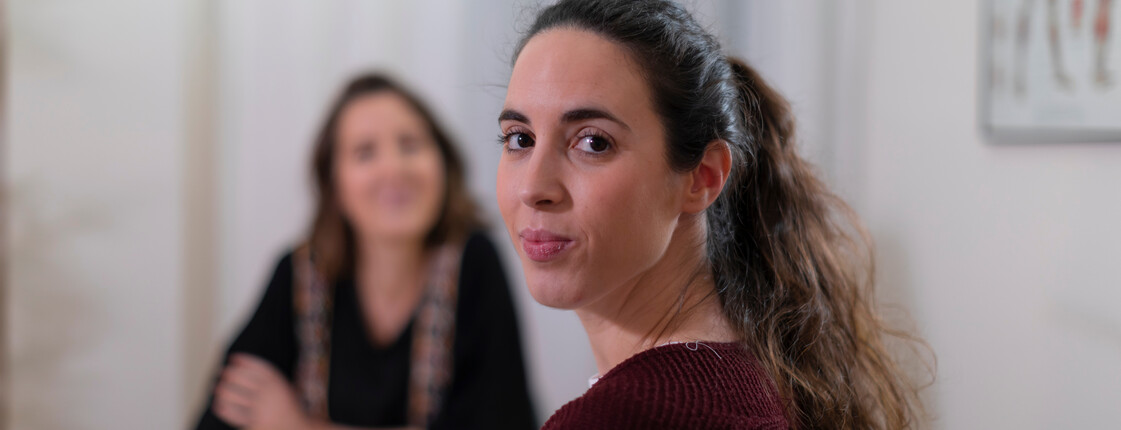 Portrait einer Frau mit dunklen Haaren, die über ihre Schulter in die Kamera schaut während sie sich in einem Beratungsgespräch befindet.
