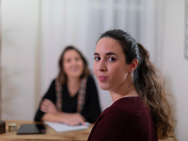 Portrait einer Frau mit dunklen Haaren, die über ihre Schulter in die Kamera schaut während sie sich in einem Beratungsgespräch befindet.