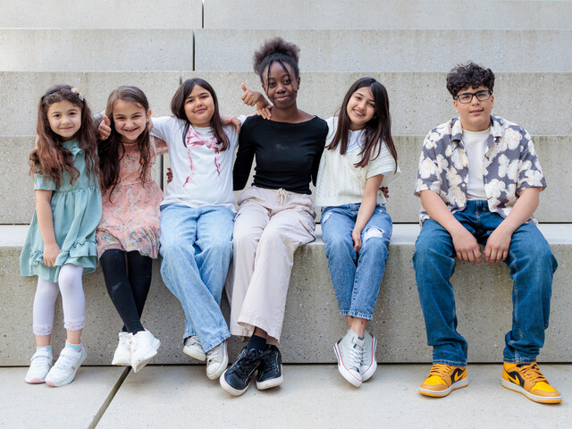 Sechs gut gelaunte Kinder sitzen nebeneinander auf einer Treppe aus Beton zu einem Gruppenfoto zusammen.