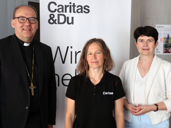 Zwei Frauen und ein Mann stehen nebeneinander vor einem Caritas Roll-Up zu einem Gruppenfoto zusammen.