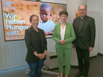 Drei Personen stehen vor einem Bildschirm mit Kampagnensujet zu einem Pressefoto zusammen.