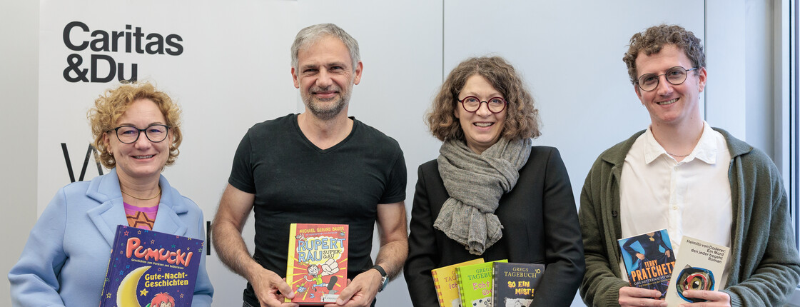 Vier Personen stehen vor einem Banner mit Caritas Logo und halten Bücher in ihren Händen.