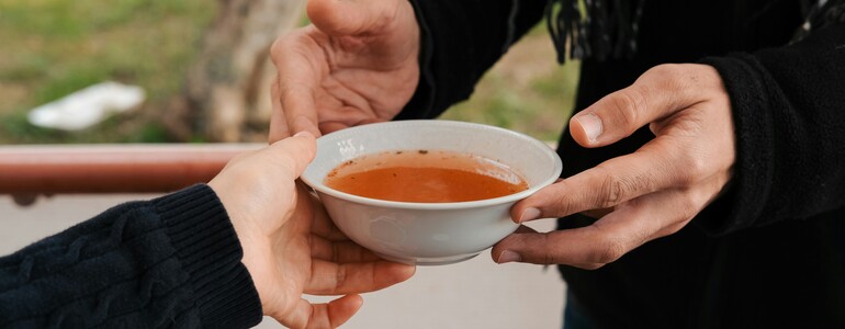 Eine Person mit schwarzer Kleidung nimmt einen Teller Suppe entgegen, der ihr gereicht wird.