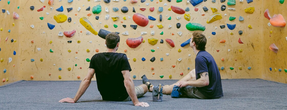 Zwei Männer sitzen in einer Kletterhalle auf dem Boden und begutachten eine Kletterwand aus Holz, an der viele bunte Klettergriffe befestigt sind.