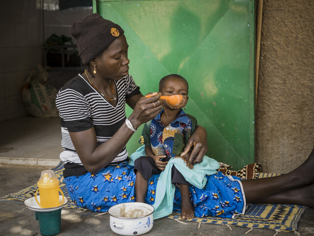Eine Mutter sitzt auf dem Boden eines Ernährungszentrums in Burkina Faso und füttert ihr Kind mit Aufbaunahrung.