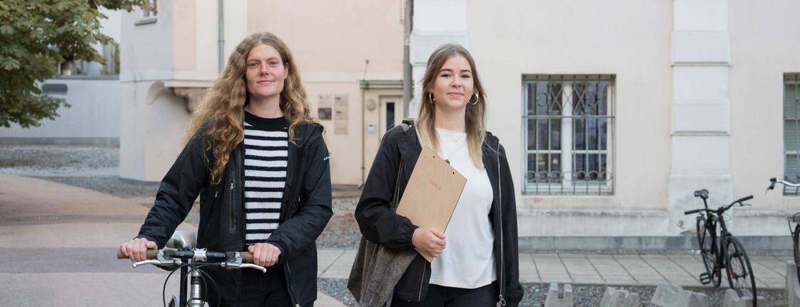 Eine Frau, die ein Fahrrad schiebt, und eine Frau mit Stoffbeutel und Klemmbrett unter ihrem Arm, laufen nebeneinander auf die kamera zu.