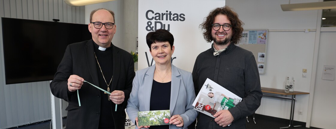 Ein Mann, eine Frau und noch ein Mann stehen nebeneinander und lachen in die Kamera. In den Händen halten sie Broschüren.