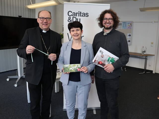 Ein Mann, eine Frau und noch ein Mann stehen nebeneinander und lachen in die Kamera. In den Händen halten sie Broschüren.