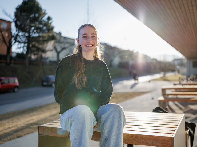 Portrait einer lachenden Frau, die vor einem Sonnenuntergang auf dem Tisch einer Sitzgruppe sitzt.